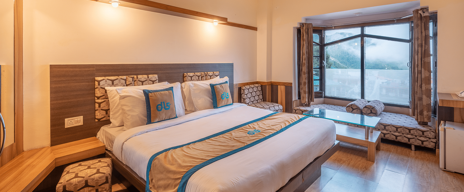 A hotel room with a large bed, a window overlooking a mountain view, and a seating area - DLS Meghavan Resort